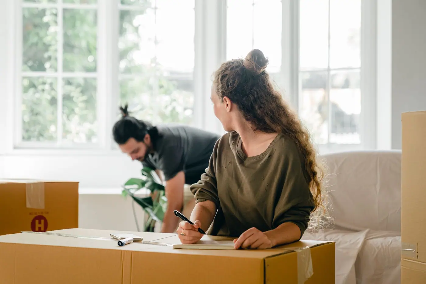 A person writing on a cardboard carton while another person moves items near a sofa
